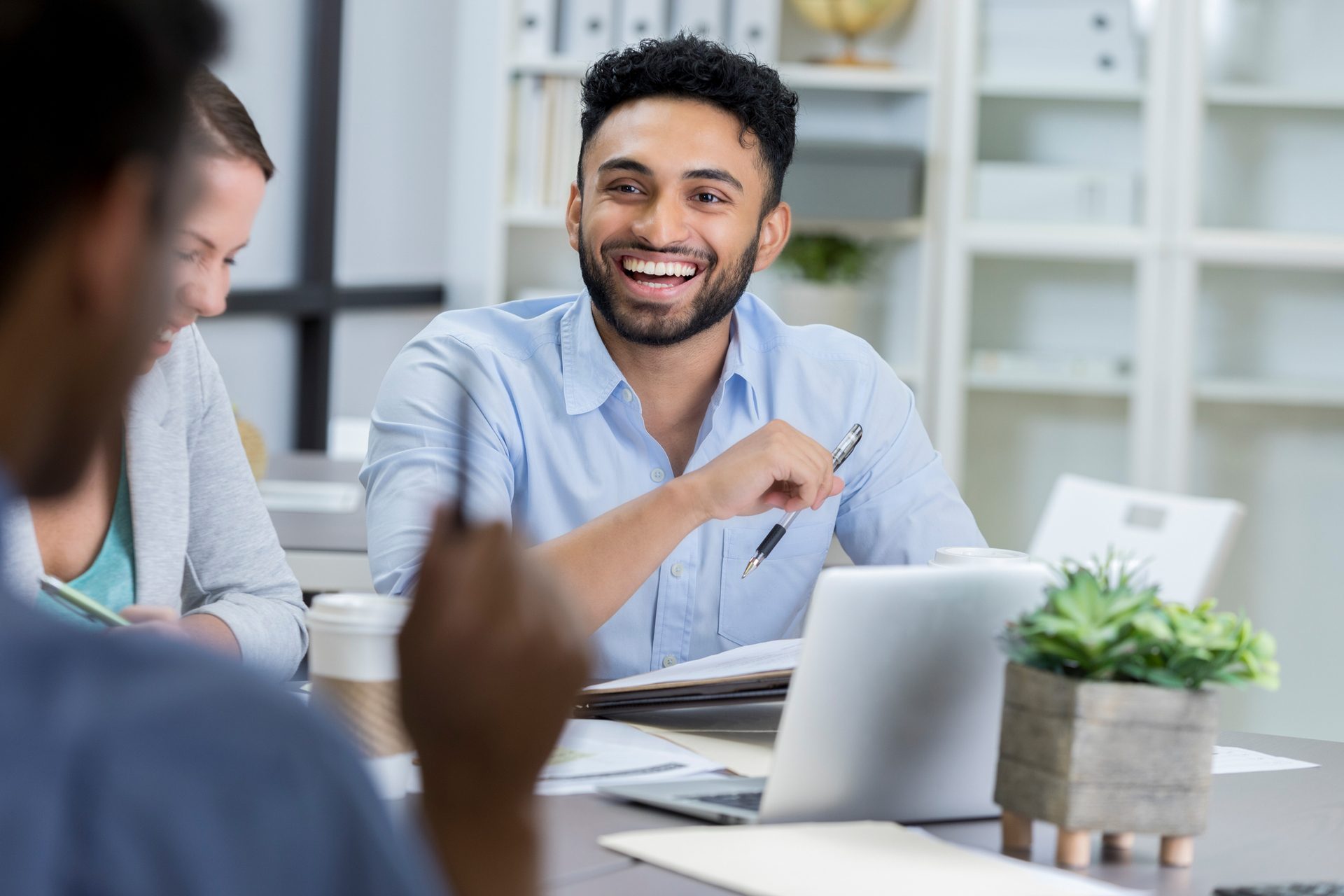 رجلٌ يرتدي قميصًا لونه أزرق فاتح يضحك مع زملائه في المكتب