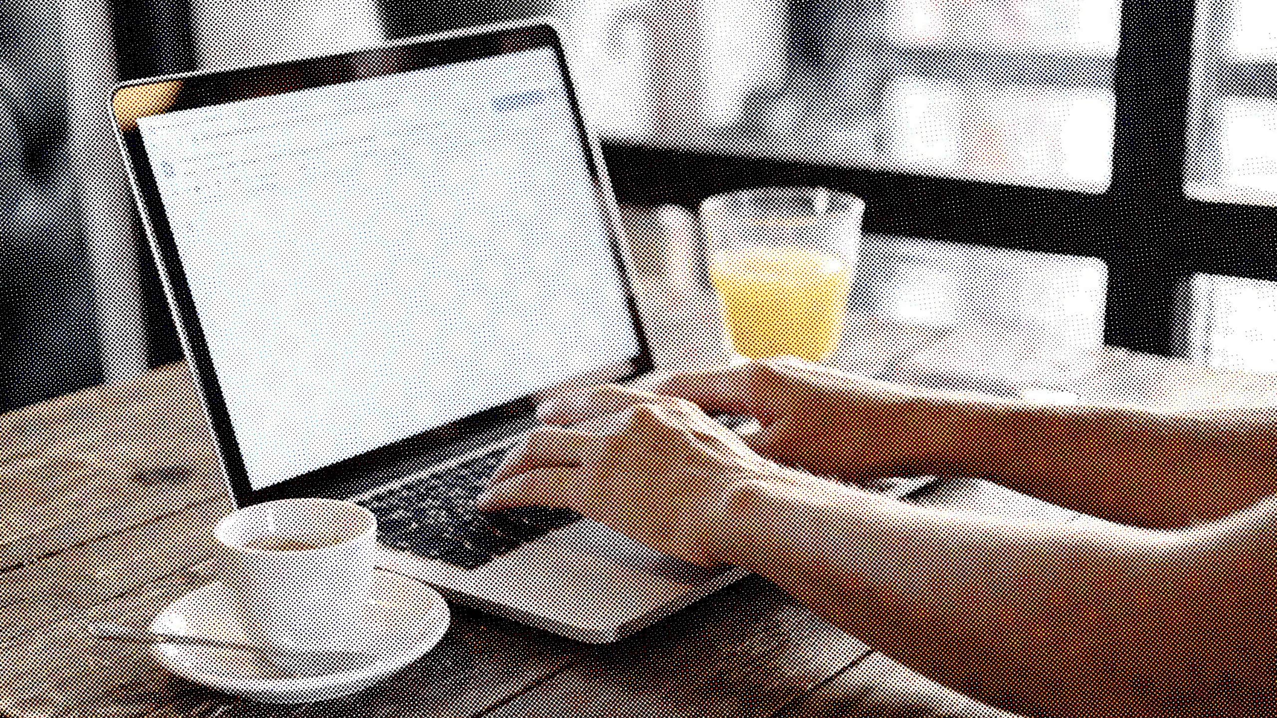 Une paire de mains qui tape sur un ordinateur portable posé sur une table en bois, à côté d'une tasse de café et d'un verre de jus d'orange